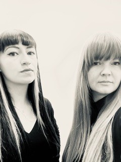 Black and white portrait of the artists.  The artists are stood together, both looking towards the camera.  They are both dressed in black and have long straight hair with fringes. 