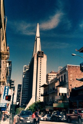 photograph of a street, the sky is blue and there is a pyramid shaped bulding in the centre of the image, surrounded by other building. People and cars can be seen in the foreground on the street. 