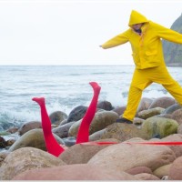 two figures on a beach, one is standing in a yellow outfit, and a second in lying down with their legs in the air wearing red tights.